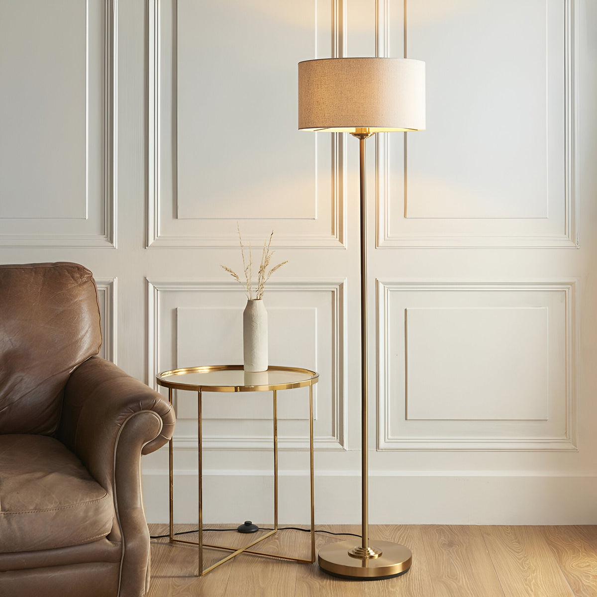 Brenley Floor Lamp in brass with a cream fabric drum shade, shown beside a leather armchair and gold side table in a living room.