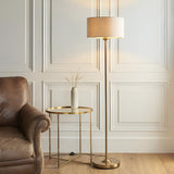 Brenley Floor Lamp in brass with a cream fabric drum shade, shown beside a leather armchair and gold side table in a living room.