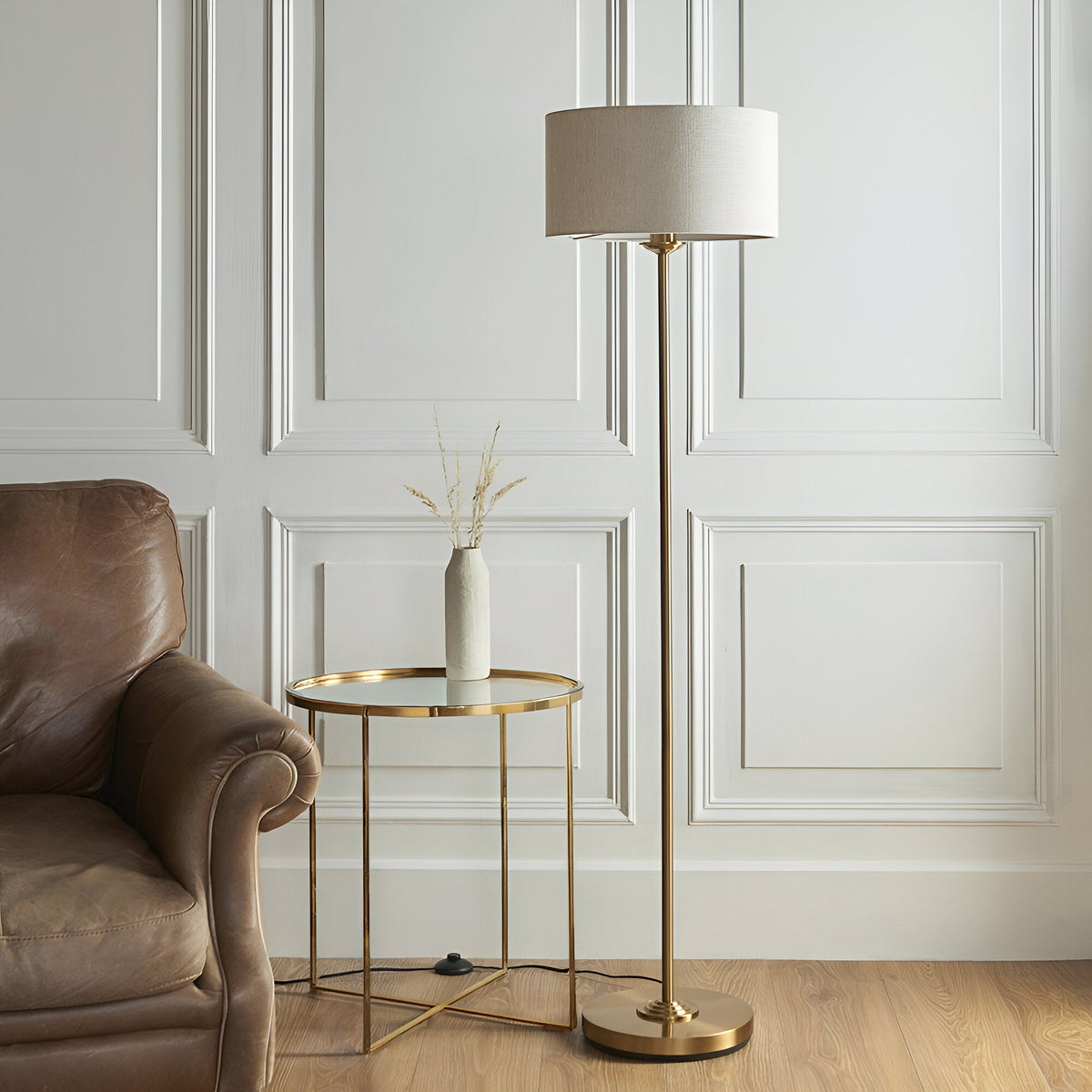Brenley Floor Lamp in brass with a round base, slim stem, and drum-shaped fabric shade, shown beside a leather armchair and gold side table.