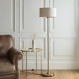 Brenley Floor Lamp in brass with a round base, slim stem, and drum-shaped fabric shade, shown beside a leather armchair and gold side table.
