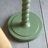 Close-up of the Brenleigh Floor Lamp's base in green, showing stacked metal spheres and a round tiered plinth on a wooden floor.
