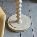 Endon Brenleigh floor lamp base in glossy white with stacked metal spheres on a round base, shown on grey plank flooring beside a yellow chair.