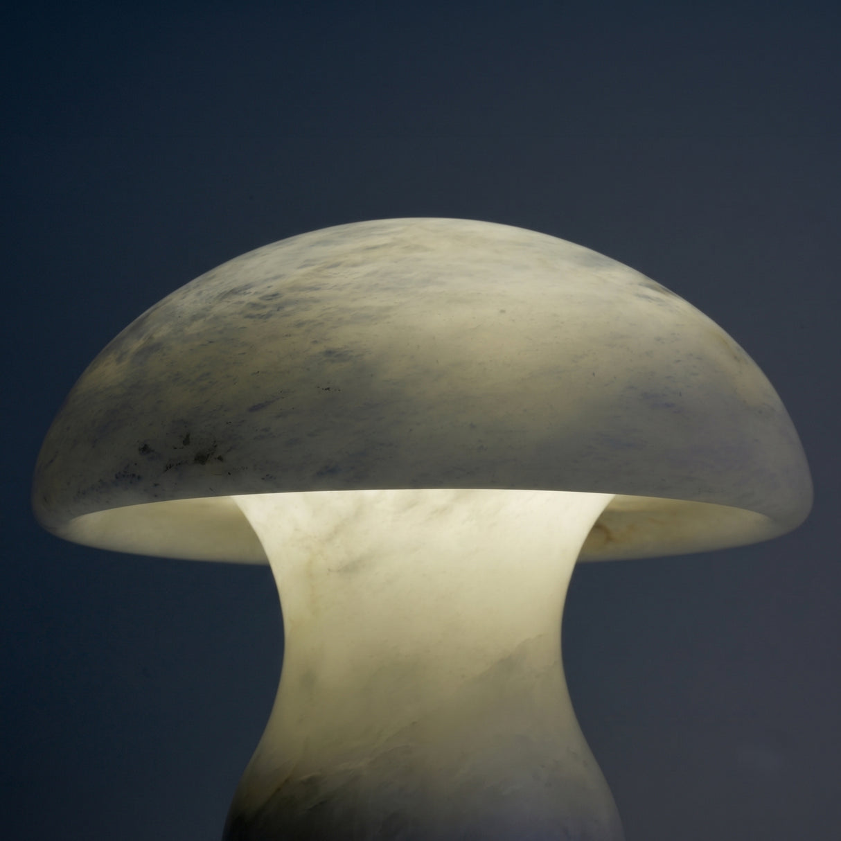 Morfia mushroom-shaped table lamp in natural alabaster, showing the dome shade lit from within with soft veining detail against a dark background.