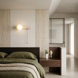 A modern, minimal bedroom with a dark wood bed, green bedding, and matching nightstand displays a potted plant and glass decor. Above the bed, the Lunaro 1 Light Bar Travertine Wall Light With Pull Switch highlights textured light beige walls.