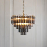 Endon Cranshaw 12 light tiered pendant with smokey grey cylindrical glass on a satin brass frame, suspended by chain against a pale panelled wall.