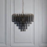 Endon Cranshaw 12 Light Tiered Pendant in satin brass with smokey grey cylindrical glass tubes, hung from a chain against a light grey panelled wall.