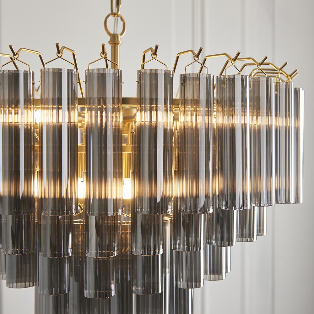 Endon Cranshaw 12 light tiered pendant with cylindrical smokey grey glass tubes on a satin brass frame, illuminated and shown against a pale wall.