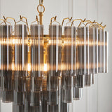 Endon Cranshaw 12 light tiered pendant with cylindrical smokey grey glass tubes on a satin brass frame, illuminated and shown against a pale wall.