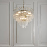 Cranshaw 12 Light Tiered Pendant in brass, with clear ribbed glass rods layered around the frame, hung from a chain against a panelled white wall.