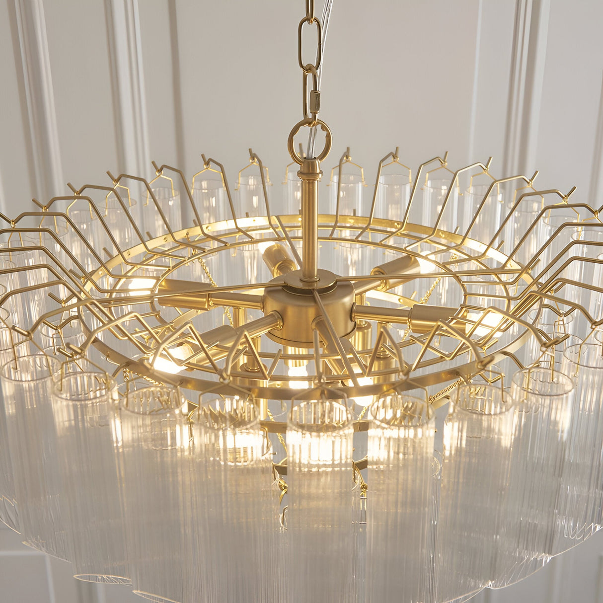 Cranshaw 12 Light Tiered Pendant in satin brass, with radiating arms and clear ribbed glass rods, suspended from a chain, shown against a light panelled wall.