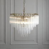Endon Cranshaw 12 light tiered pendant in satin brass with ribbed clear glass cylinders, suspended from a chain against a panelled grey wall.