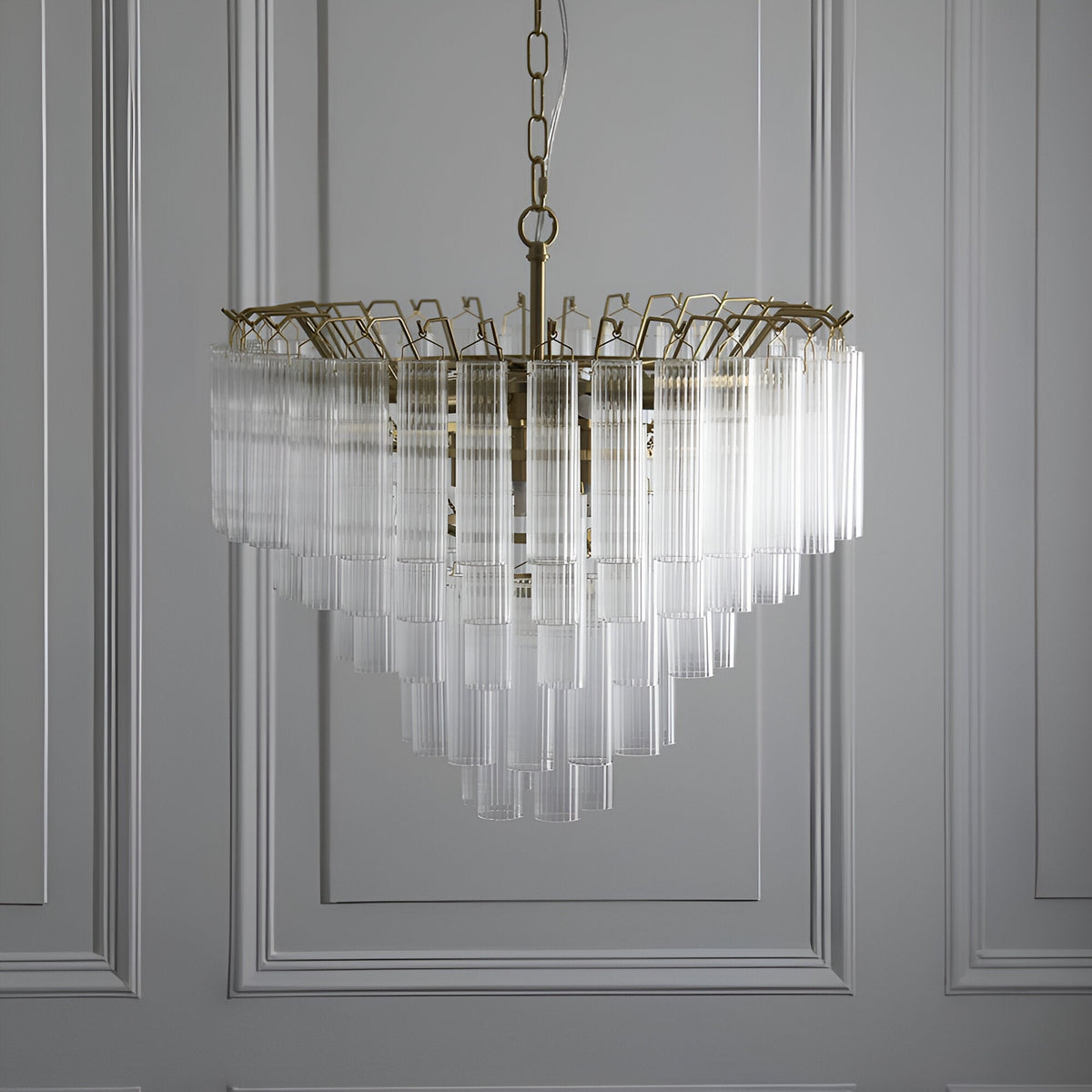 Endon Cranshaw 12 light tiered pendant in satin brass with ribbed clear glass cylinders, suspended from a chain against a panelled grey wall.