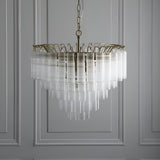 Endon Cranshaw 12 light tiered pendant in satin brass with ribbed clear glass cylinders, suspended from a chain against a panelled grey wall.