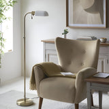 Endon Chartbourne floor lamp with slim antique brass stem and gloss opal glass shade, beside a beige wingback armchair in a bright living room.