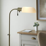 Endon Chartbourne floor lamp with antique brass curved stem and gloss opal glass shade, shown beside a wooden desk in a bedroom setting.