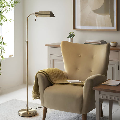 Endon Chartbourne floor lamp with slim antique brass stem and angled metal shade, beside a mustard velvet armchair in a bright living room.