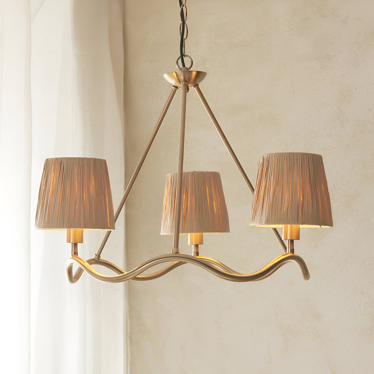 Danemont 3 Light Pendant in brass with a wave-shaped hoop and three hand-wrapped raffia shades, hung on a chain against a neutral wall.