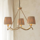 Danemont 3 Light Pendant in brass with a wave-shaped hoop and three hand-wrapped raffia shades, hung on a chain against a neutral wall.