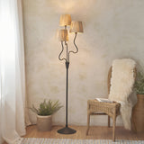 Endon Danemont 3 light floor lamp in matt black with wavy stems and hand-wrapped raffia tapered shades, styled in a boho corner beside a rattan chair.