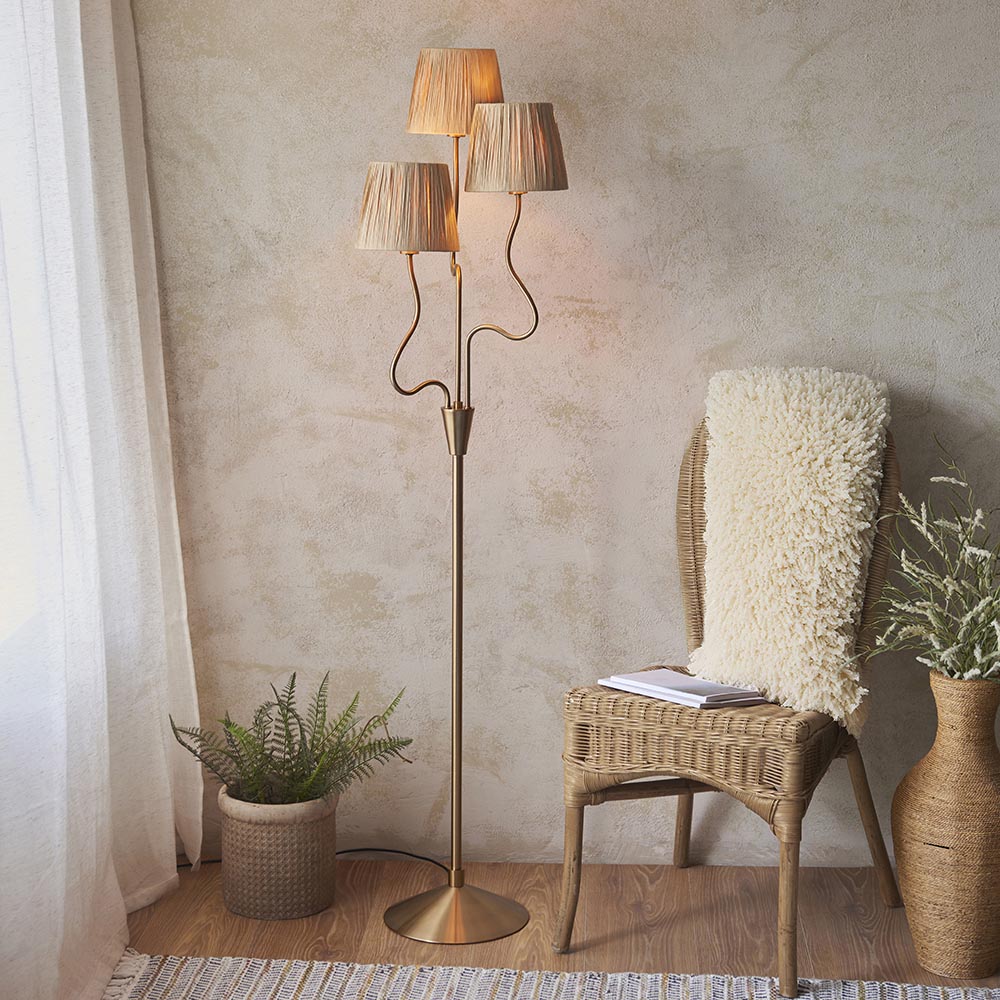 Danemont 3 Light Floor Lamp in aged brass with wriggle stem arms and tapered raffia shades, shown beside a wicker chair in a living room.