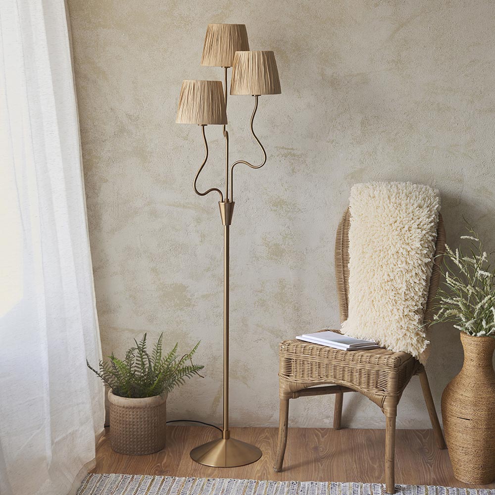 Danemont 3 Light Floor Lamp in aged brass with wriggle stem arms and tapered raffia shades, shown beside a rattan chair and plants.
