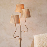 Danemont 3 Light Floor Lamp in aged brass with wriggle stem and three tapered raffia shades, shown in a room with a woven chair and cushion.