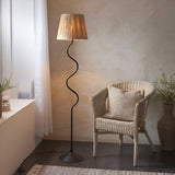 Endon Danemont floor lamp with matt black wriggle-shaped metal stem and tapered hand-wrapped raffia shade, beside a rattan chair in a neutral corner.