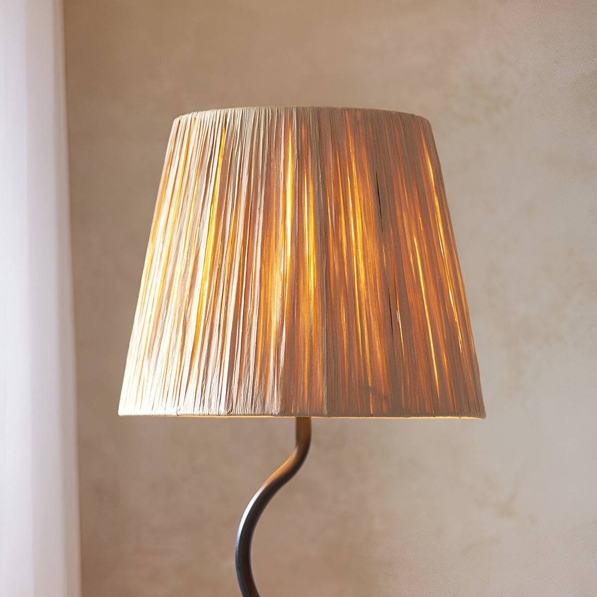 Endon Danemont floor lamp illuminated, showing tapered hand-wrapped raffia shade glowing warmly above a matt black wriggle-shaped stem against a neutral wall.