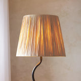 Endon Danemont floor lamp illuminated, showing tapered hand-wrapped raffia shade glowing warmly above a matt black wriggle-shaped stem against a neutral wall.