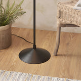 Close-up of the matt black round metal base and stem of the Endon Danemont floor lamp on wood flooring beside a rattan chair and potted fern.