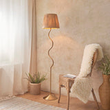 Endon Danemont floor lamp in aged brass with a wriggle-shaped stem and tapered raffia shade, styled beside a rattan chair and fern in a warm neutral room.