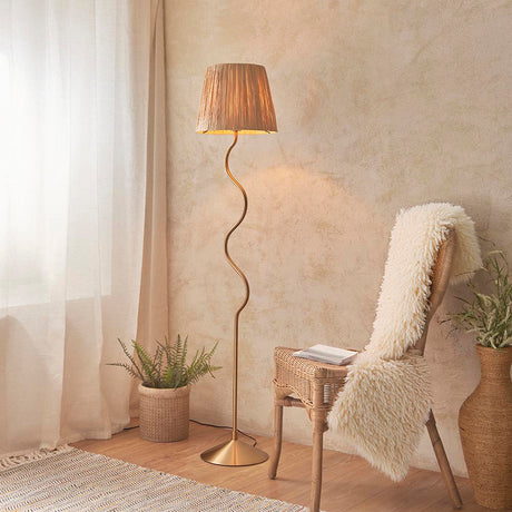 Endon Danemont floor lamp in aged brass with a wriggle-shaped stem and tapered raffia shade, styled beside a rattan chair and fern in a warm neutral room.