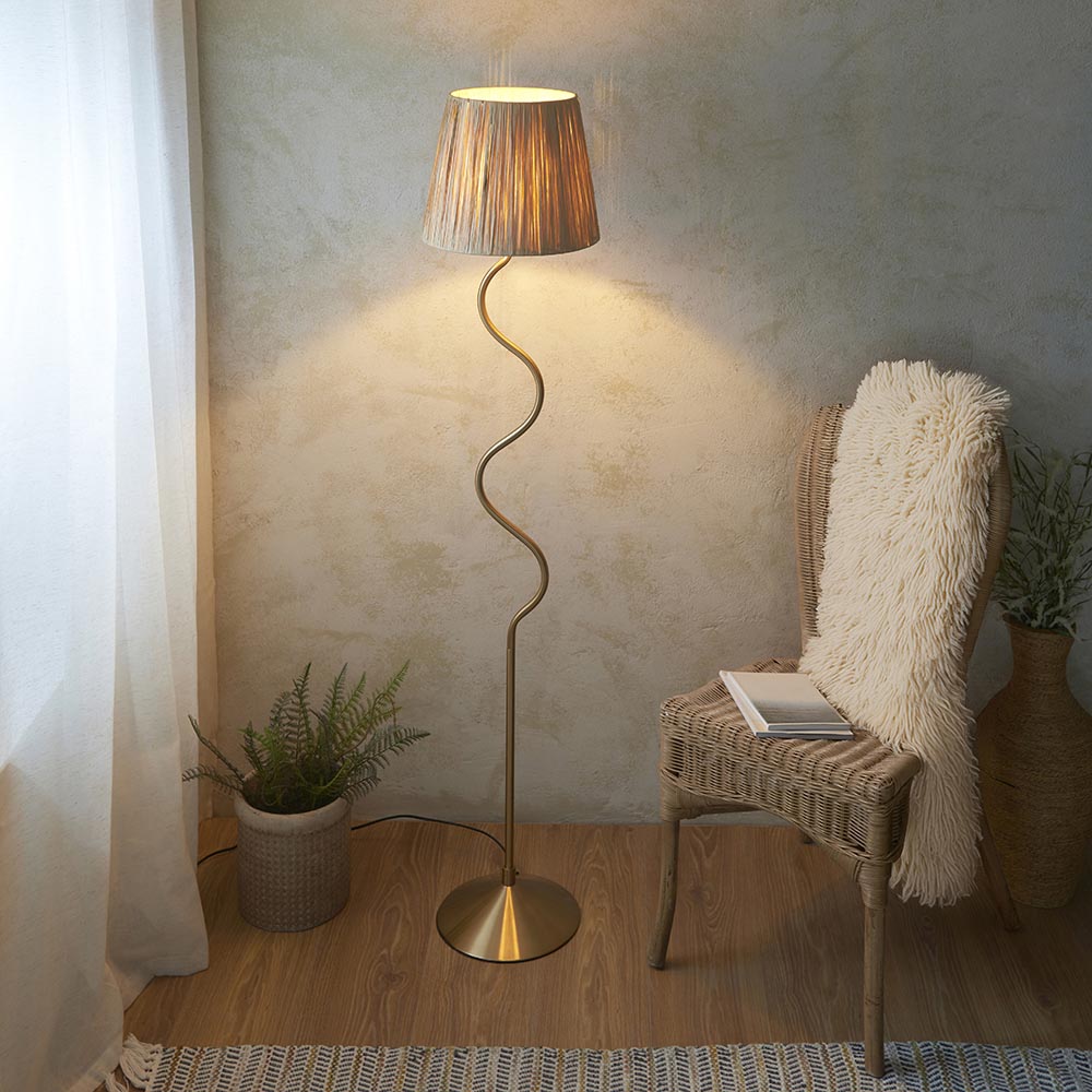 Endon Danemont floor lamp in aged brushed brass with a wriggle stem and tapered raffia shade, lit beside a rattan chair, fern and textured wall.