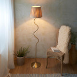 Endon Danemont floor lamp in aged brushed brass with a wriggle stem and tapered raffia shade, lit beside a rattan chair, fern and textured wall.