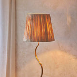 Endon Danemont floor lamp with a wriggle-shaped aged brass stem and tapered hand-wrapped raffia shade, lit beside a pale plaster wall.