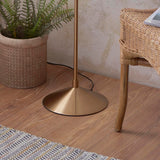 Close-up of the Endon Danemont floor lamp's aged brass tapered base and stem on wood flooring beside a rattan chair and potted fern.