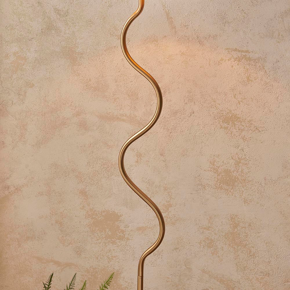 Endon Danemont floor lamp with a wriggle-shaped brushed aged brass stem, shown against a warm neutral plaster wall with a fern below.