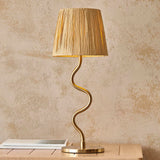 Endon Danemont table lamp with a wavy brushed brass stem and tapered natural raffia shade, on a wooden surface against a textured beige wall.