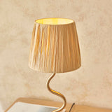 Endon Danemont table lamp lit on a wooden surface, with a tapered natural raffia shade on a wavy brushed brass stem against a neutral wall.