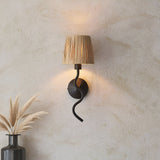 Endon Danemont wall light with wavy matt black arm and natural raffia shade, mounted on a textured cream wall beside dried pampas grass.