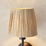 Close-up of Endon Danemont wall light shade in wrapped natural raffia with tapered form, mounted on a matt black arm against a textured cream wall.