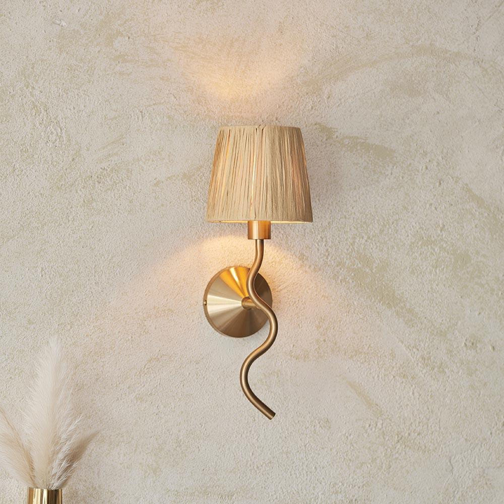 Danemont Wall Light in brass with a wriggle-shaped metal arm and natural raffia shade, mounted on a textured wall.