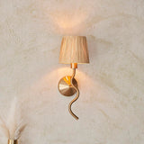 Danemont Wall Light in brass with a wriggle-shaped metal arm and natural raffia shade, mounted on a textured wall.