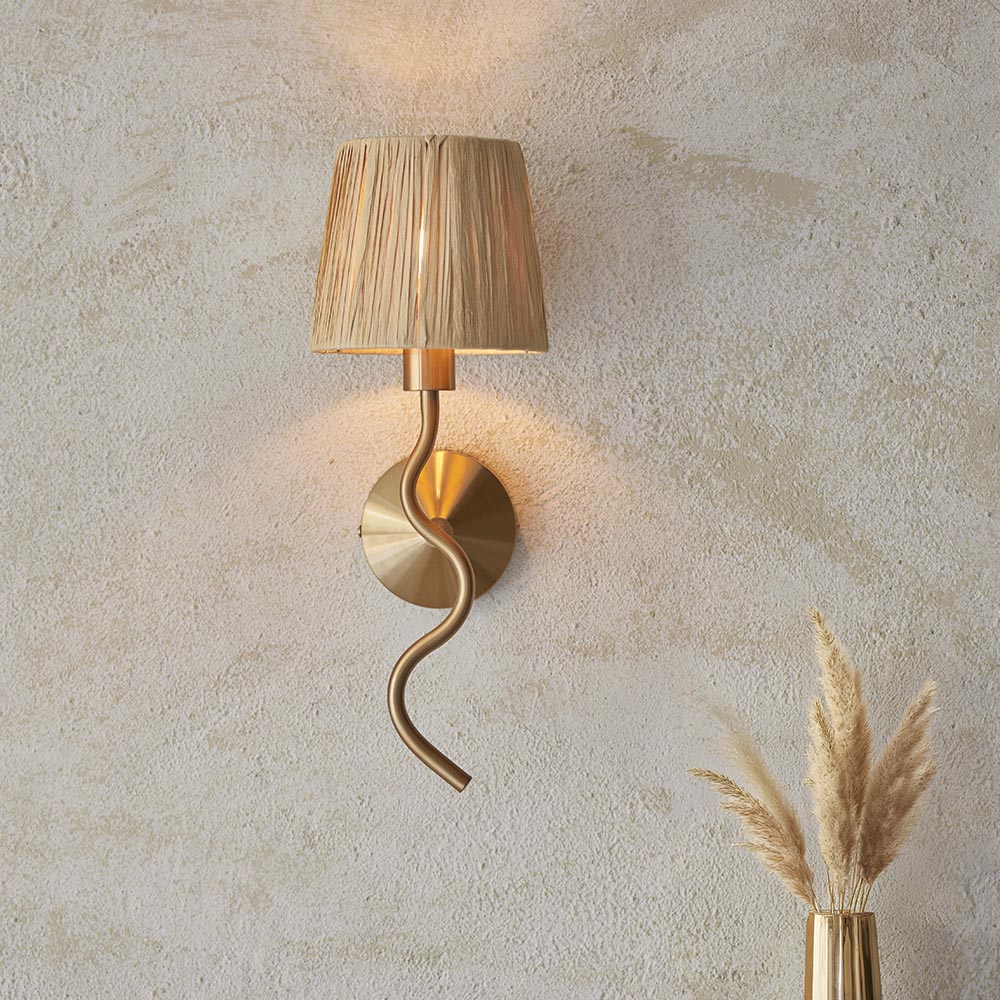Danemont Wall Light in brass with a wriggle-shaped arm and natural raffia shade, mounted on a plaster wall beside a vase of pampas grass.