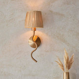 Danemont Wall Light in brass with a wriggle-shaped arm and natural raffia shade, mounted on a plaster wall beside a vase of pampas grass.
