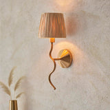 Danemont Wall Light in brass with a wriggle-shaped arm and natural raffia shade, mounted on a light textured wall beside dried pampas grass.