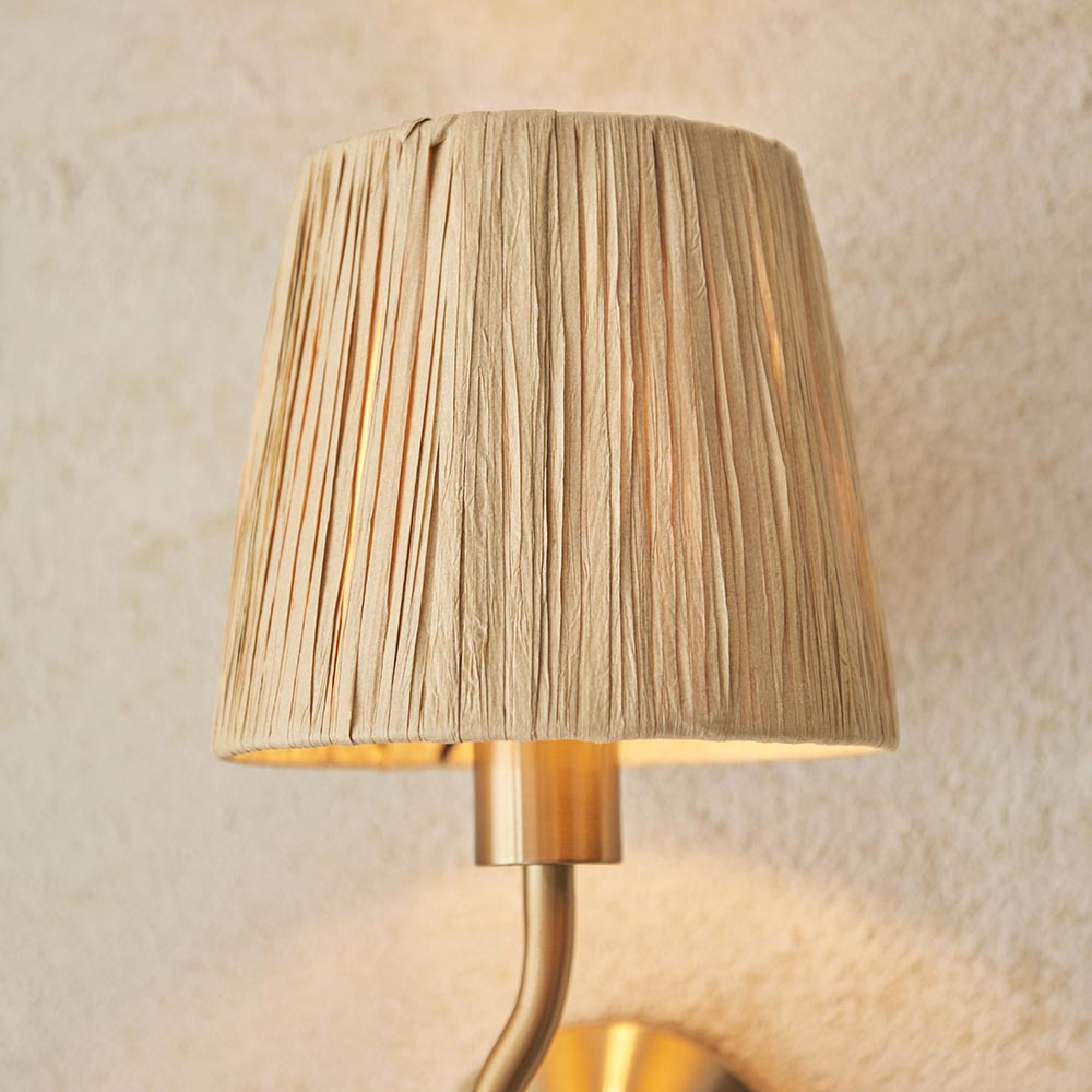 Brass Danemont wall light with curved arm and natural raffia shade, glowing warmly against a textured light background.