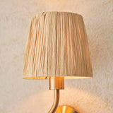 Brass Danemont wall light with curved arm and natural raffia shade, glowing warmly against a textured light background.