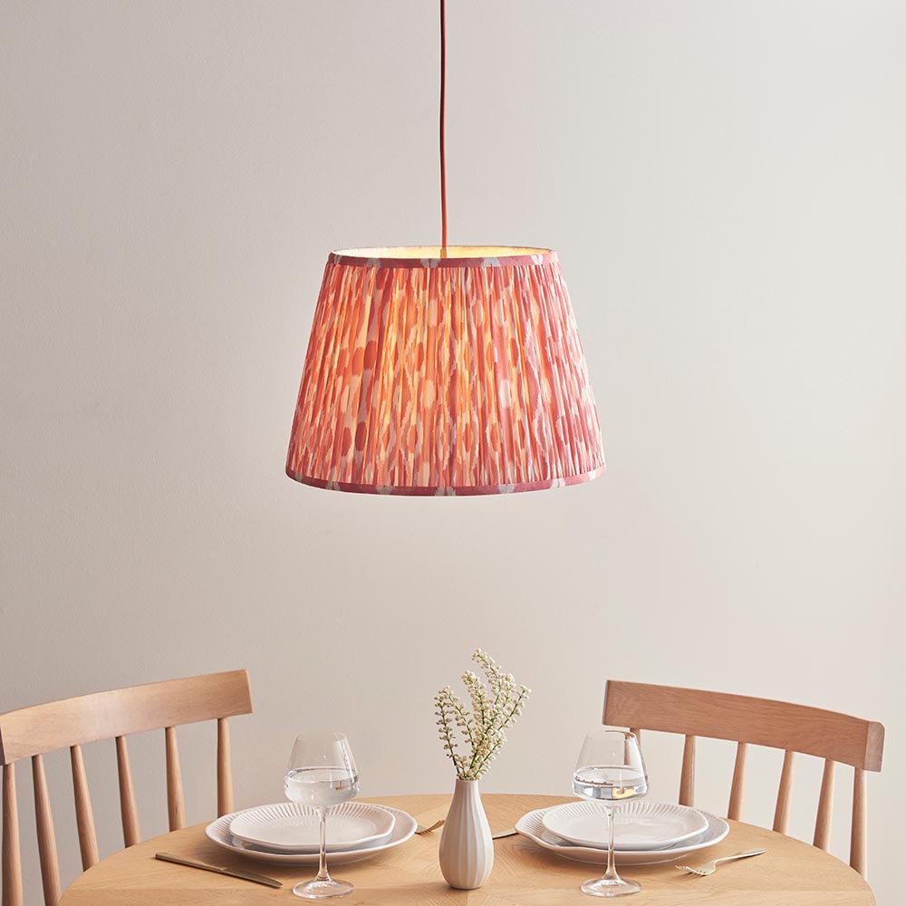 Endon Brackmont Ikat Coral pendant light with handmade pleated tapered faux silk shade in pink and red, on a red braided cable, above a wooden dining table.