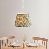 Brackmont Pendant light with a pleated green fabric tapered shade and brass fitting, hanging above a wooden dining table in a light-toned room.